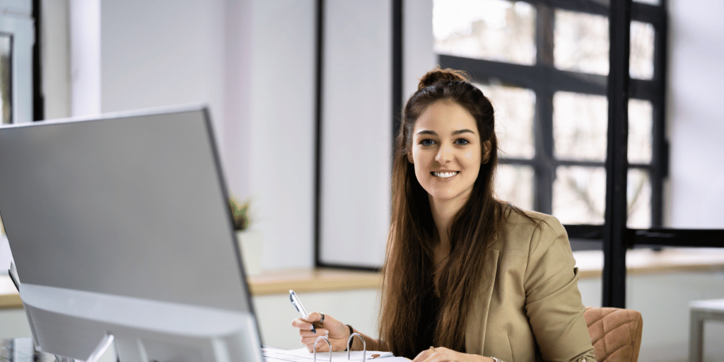 Woman working at a desktop computer with and open file and a smile for the camera.