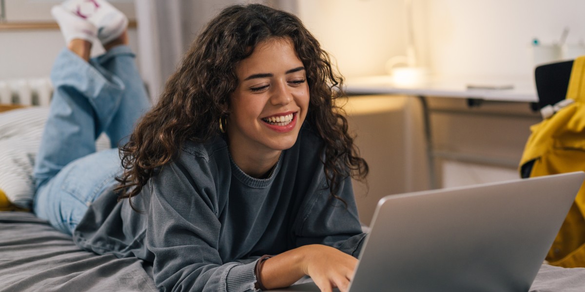 student smiling while working on her laptop, on the bed of her new student room.