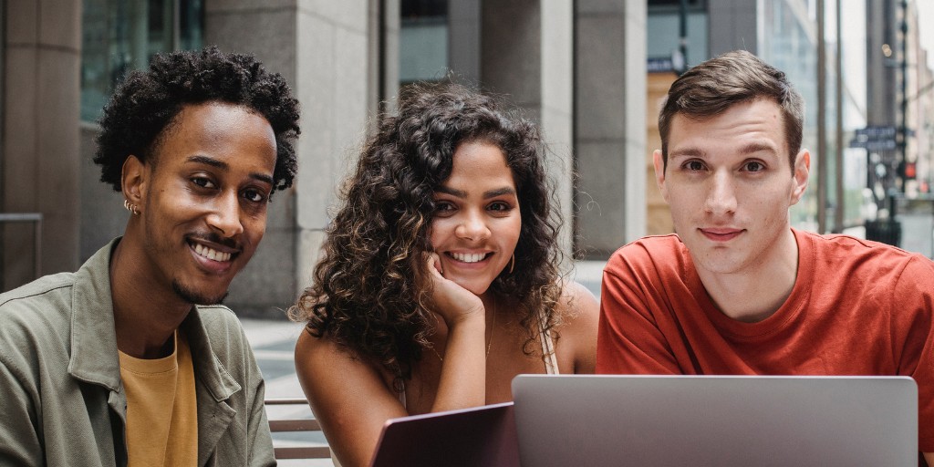 3 students smiling at the camera and sitting at a desk outside with their laptops