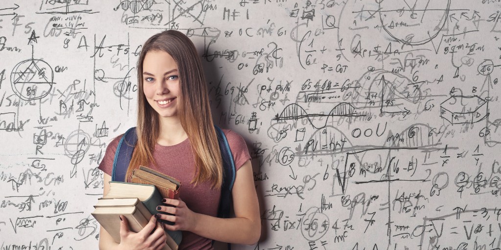 Female student holding books and smiling at the camera. Behind her is a board of mathematical workings.