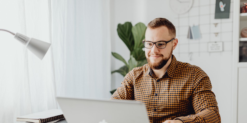 Smiling man looking at laptop while sitting at a desk. There is a plant behind him.