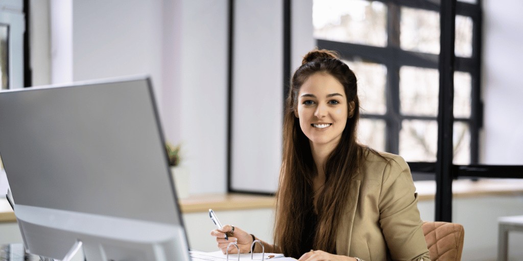 Woman working at a desktop computer with and open file and a smile for the camera.