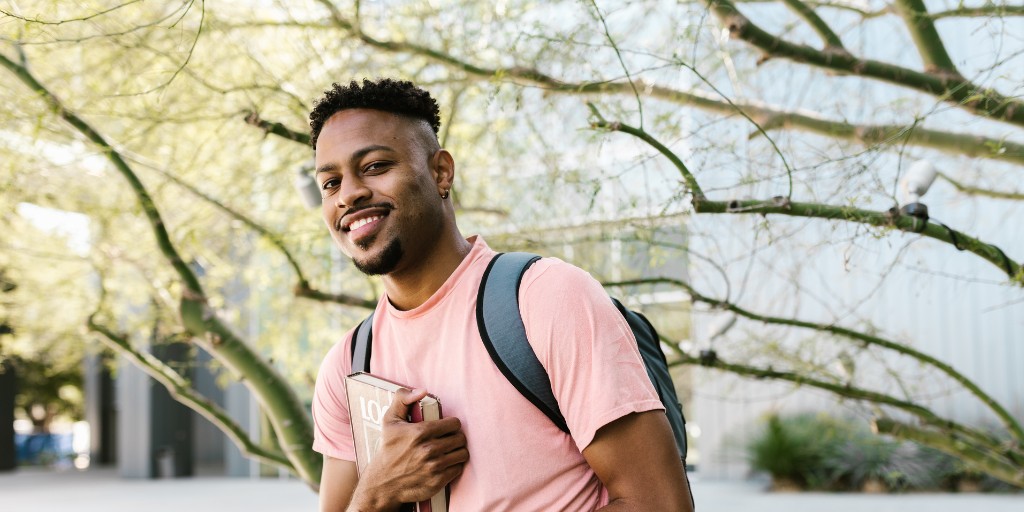 A smiling student stands outdoors holding a book, a backpack over one shoulder