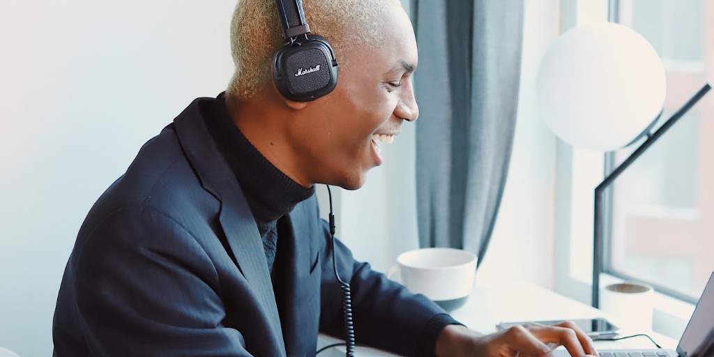 Young man in a suit, wearing headphones and laughing in conversation with someone on his laptop
