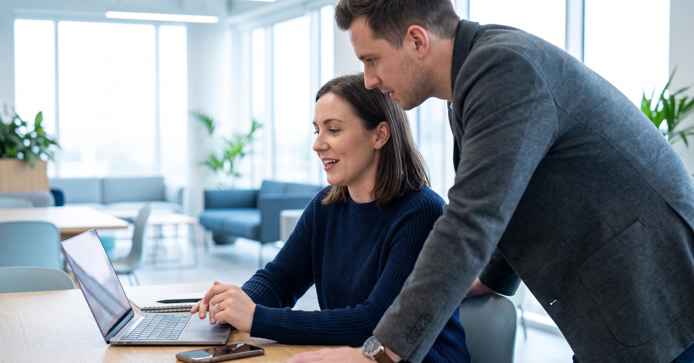 Two colleagues look at a laptop together in a bright office, one seated and one standing