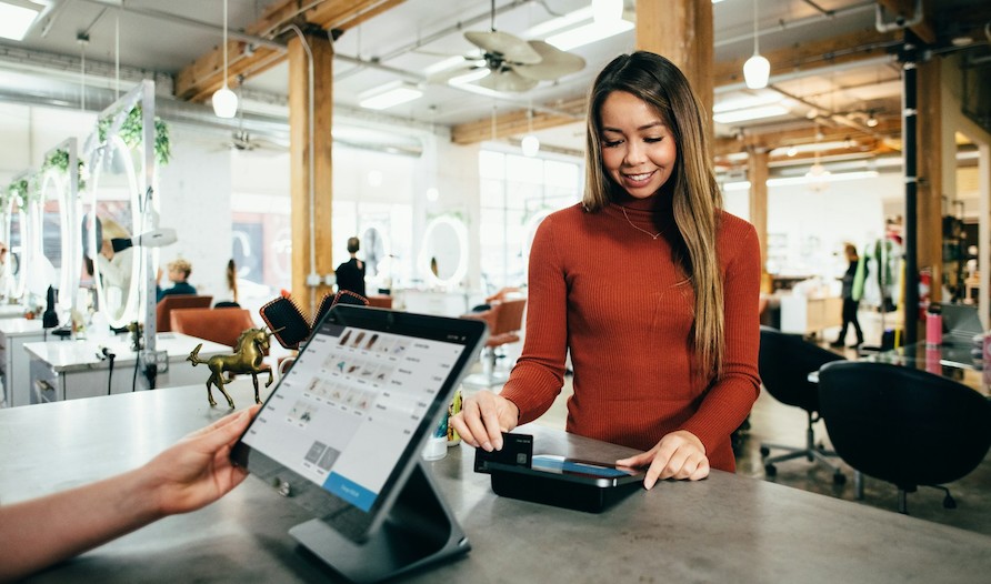 A customer taps a bank card on a payment terminal at a shop counter while a staff member holds a tablet